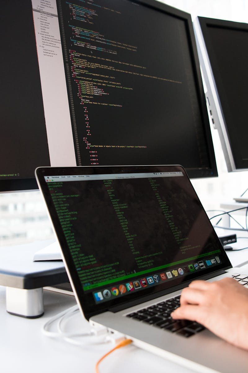 A developer writes code on a laptop in front of multiple monitors in an office setting.